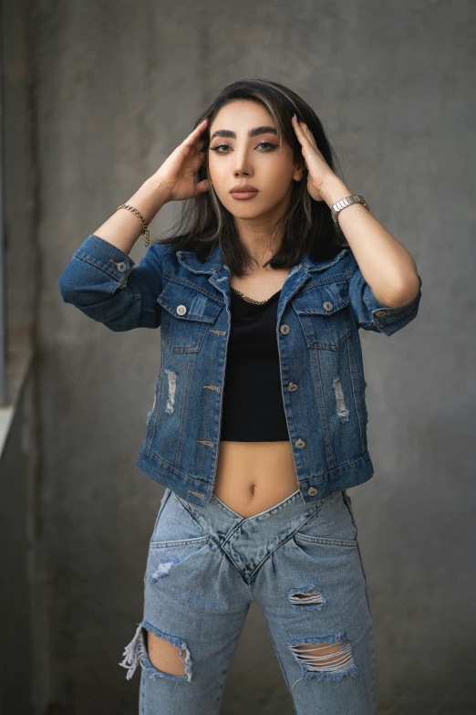 a woman in a black top and blue jean jacket stands with her hands behind her head