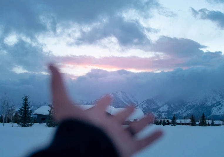 a person's hand pointing at the snow with mountains behind