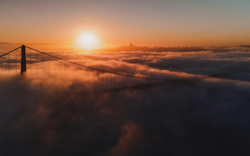 the sun is setting behind a suspension bridge with clouds and fog