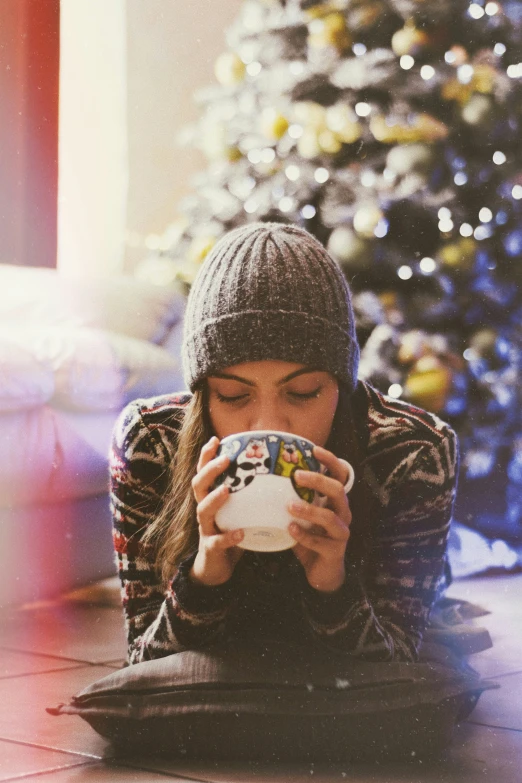 a woman is holding a mug while sitting near a christmas tree