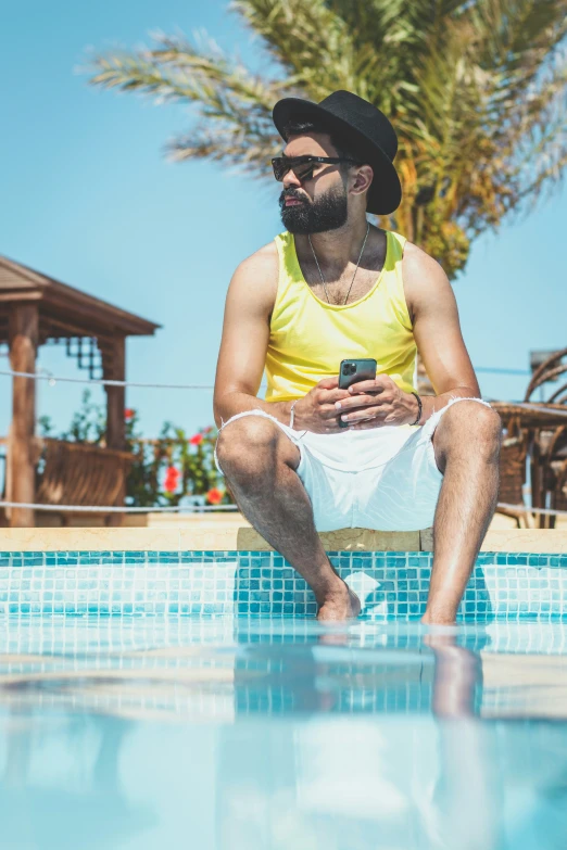 the man in the hat sits on the edge of a pool while looking at his cell phone