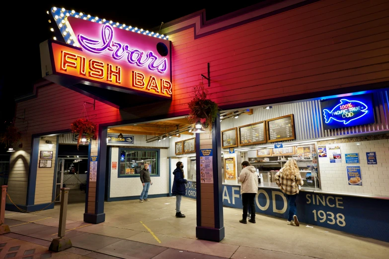 people stand outside a restaurant where the signs are lit up