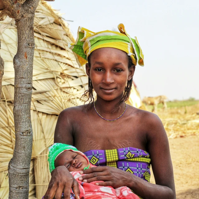 an african woman holding her baby in front of a hut