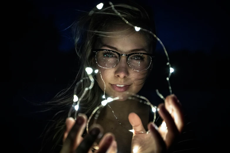 a girl wearing glasses has her hand under some string lights