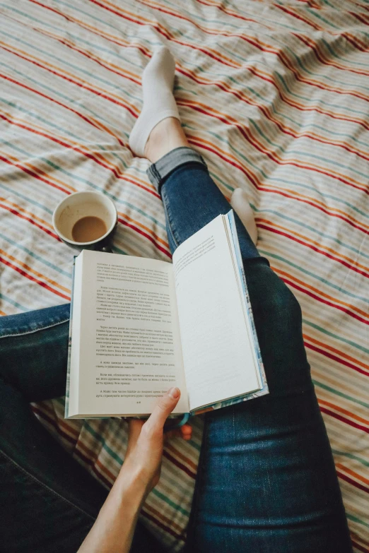 a person sits in bed holding a book with a cup of coffee
