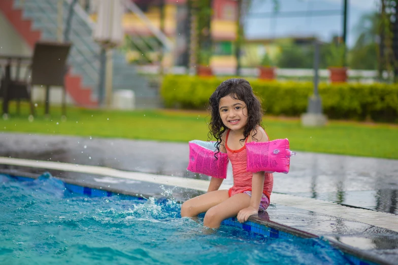 little girl in pink bathing suit sitting on a ledge by swimming pool