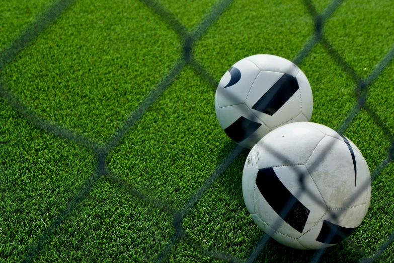 two soccer balls resting on the grass behind the fence