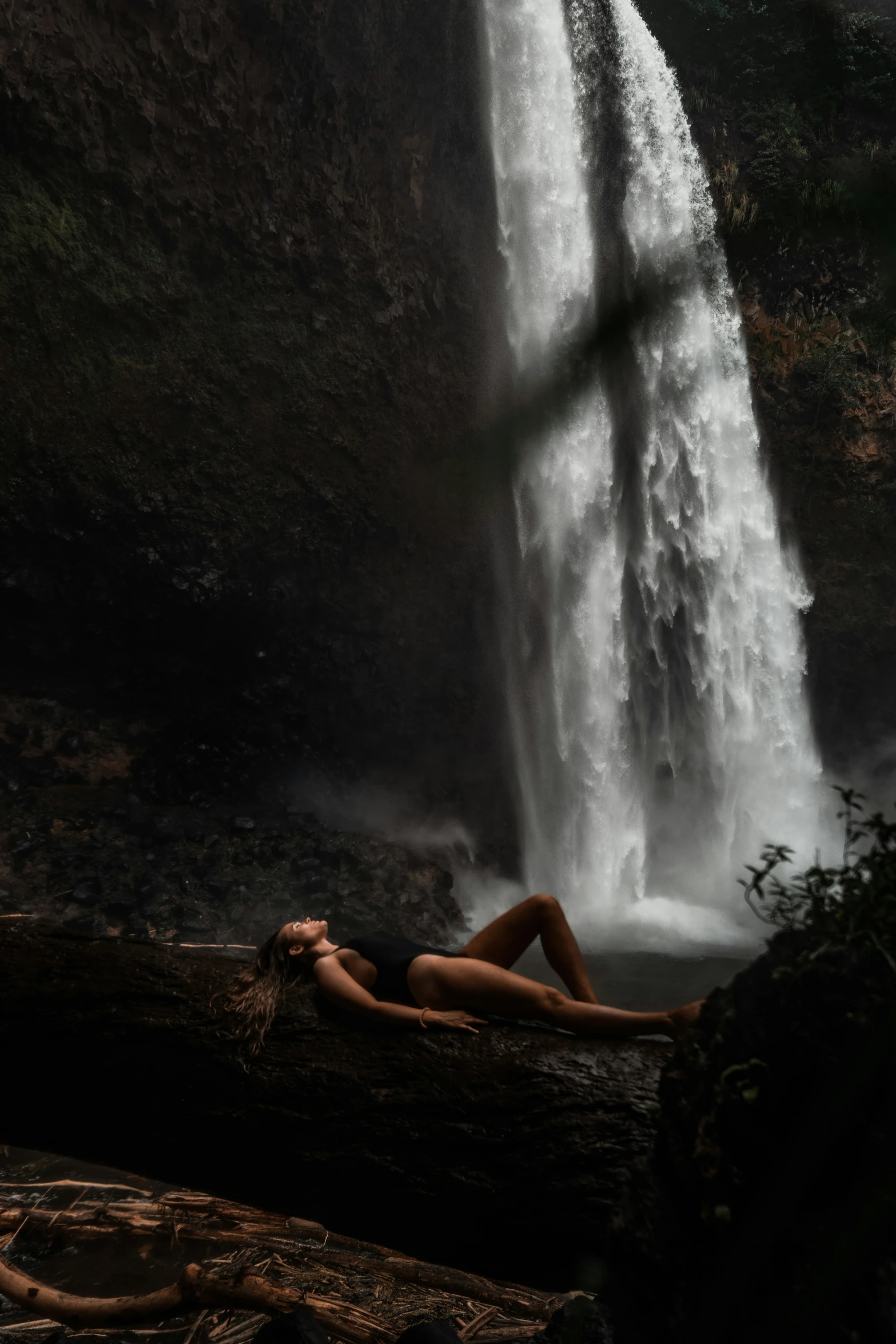 a woman in a bathing suit rests on a log below a waterfall