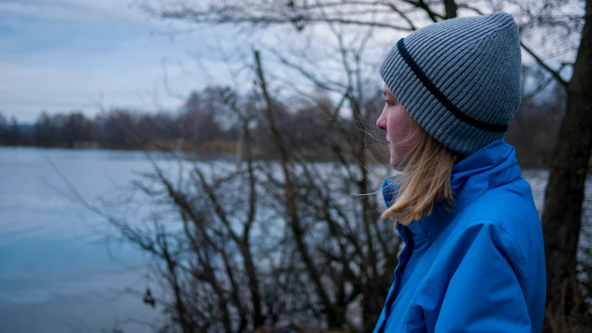 a girl in blue coat standing next to trees and water