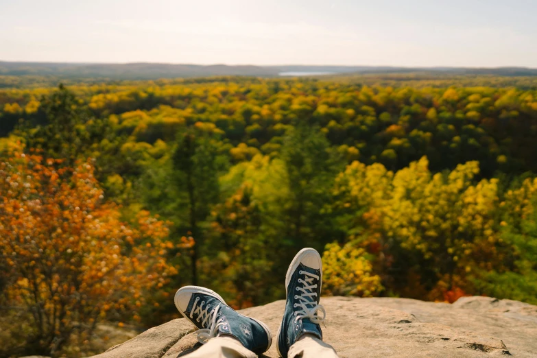 person wearing tennis shoes sitting on rock ledge overlooking forest