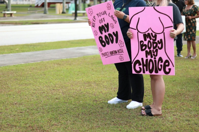 two woman holding pink posters while standing on the grass