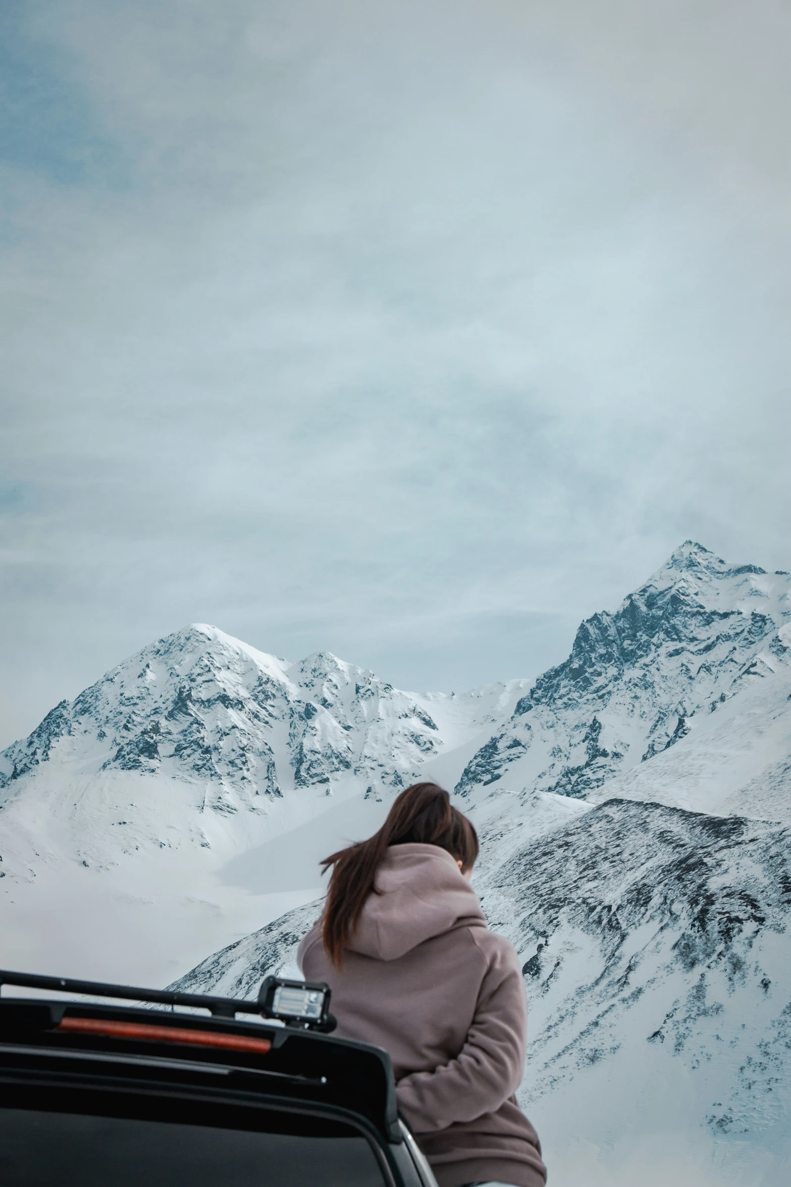 woman in hoodie sitting on roof of car looking over mountains