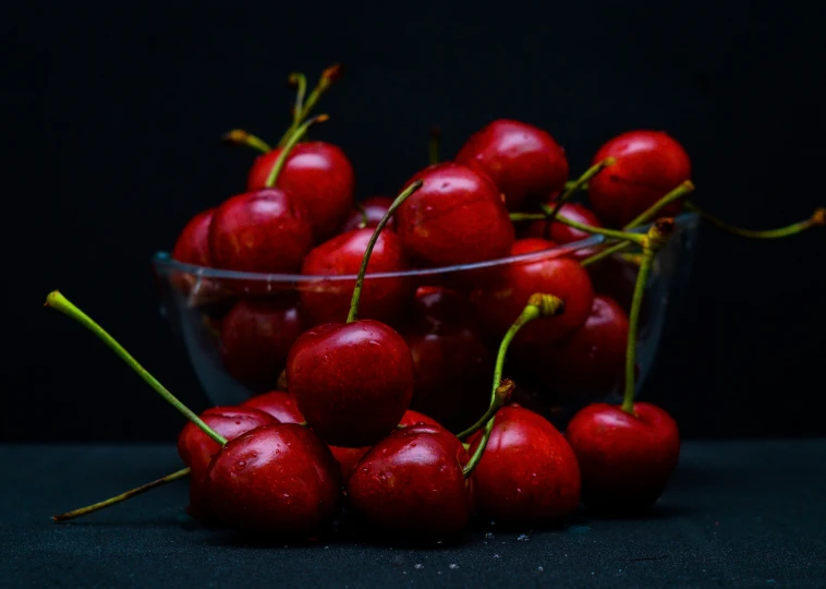 a clear bowl filled with red cherries on a dark background