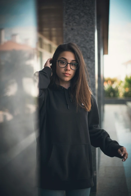 a woman in glasses and a black sweatshirt leaning against a wall