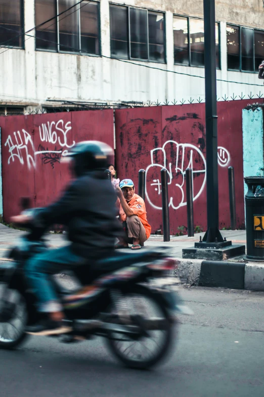 two people sitting on a motorcycle riding past a red building