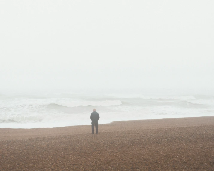 man in grey coat looking at ocean while standing on foggy beach