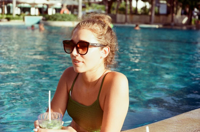 a girl sitting in front of a swimming pool holding a drink