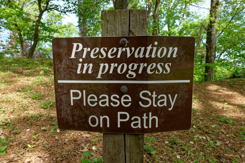 a sign reading pression un progress please stay on path is posted on the side of a tree in a wooded area