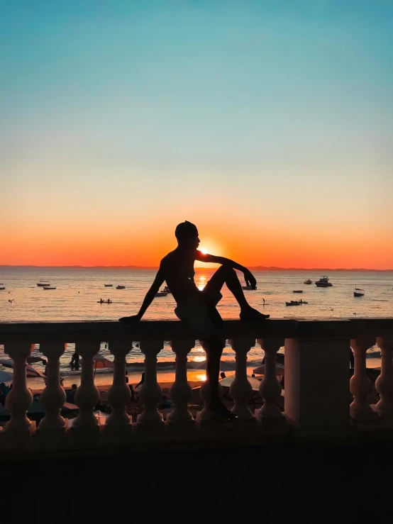 man sitting on railing with surfboard watching the sun go down