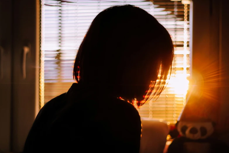 a girl stares through the blinds at the light coming through