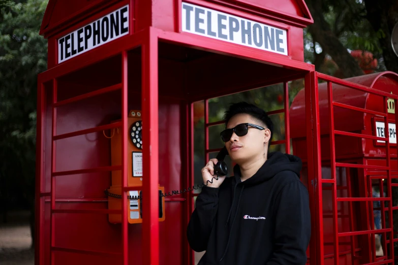 young man in black hoodie and shades standing by red phone booth