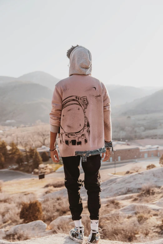 man wearing pink sweatshirt and black pants and white sneakers stands looking out over rocky hills
