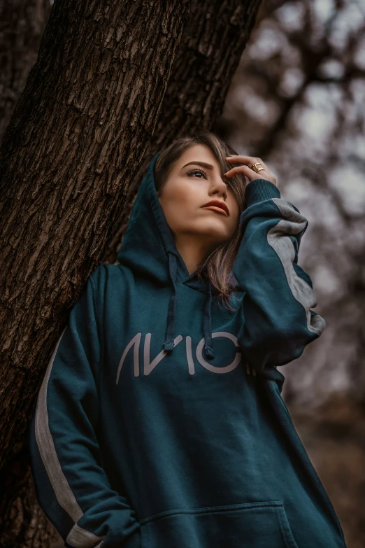 a woman wearing a green sweatshirt and hoodie leaning against a tree