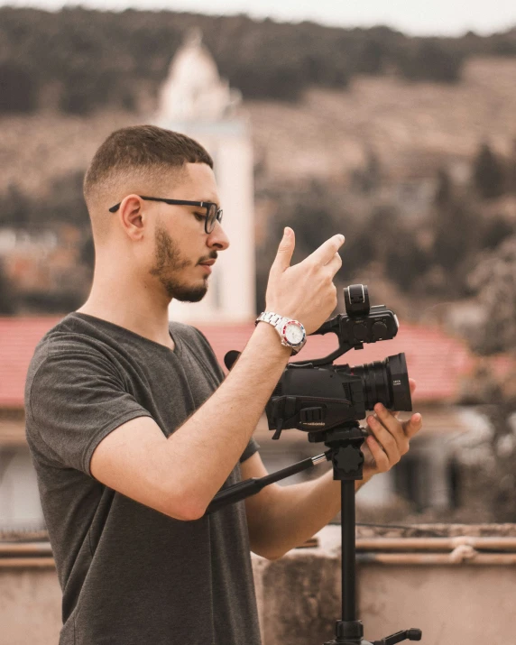 a man in glasses and t - shirt holds a camera and stands behind an on tripod