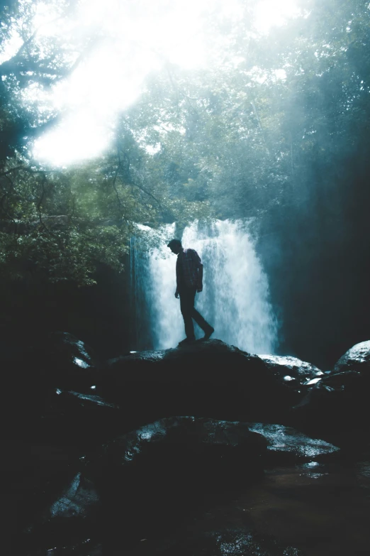 the silhouette of a man standing by the water below a waterfall