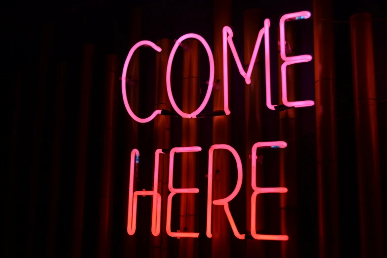 the words'come here'neon in bright pink against a dark backdrop