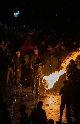 a crowd of people stand around a bonfire with it's light on