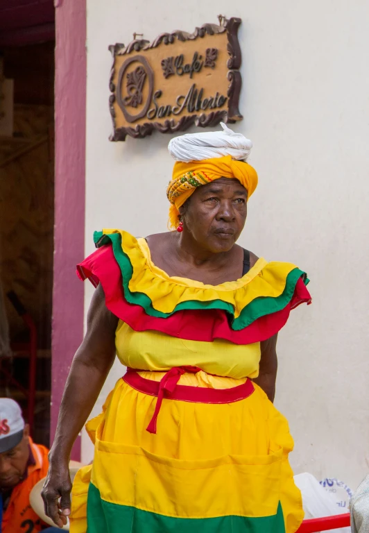 an older woman in brightly colored attire stands by a white wall