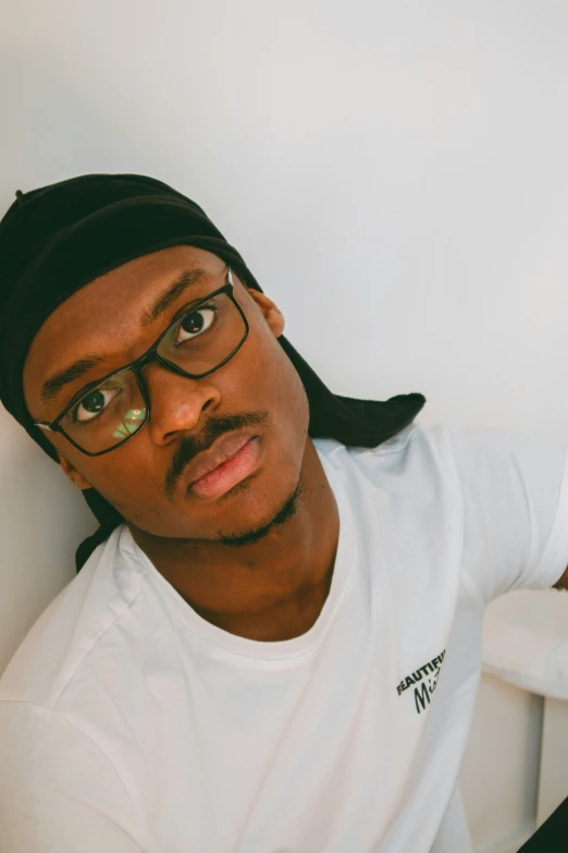 a man with glasses and a black hat leaning against a wall