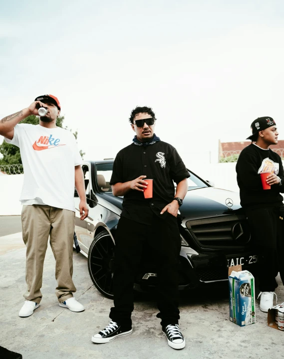three men with beverages in their hands standing by a parked car