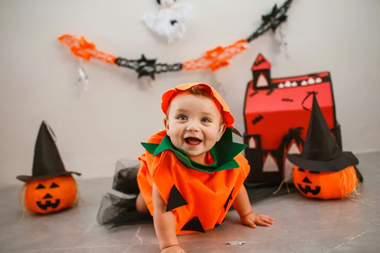 a baby girl wearing a pumpkin outfit sitting on the floor with other halloween decorations