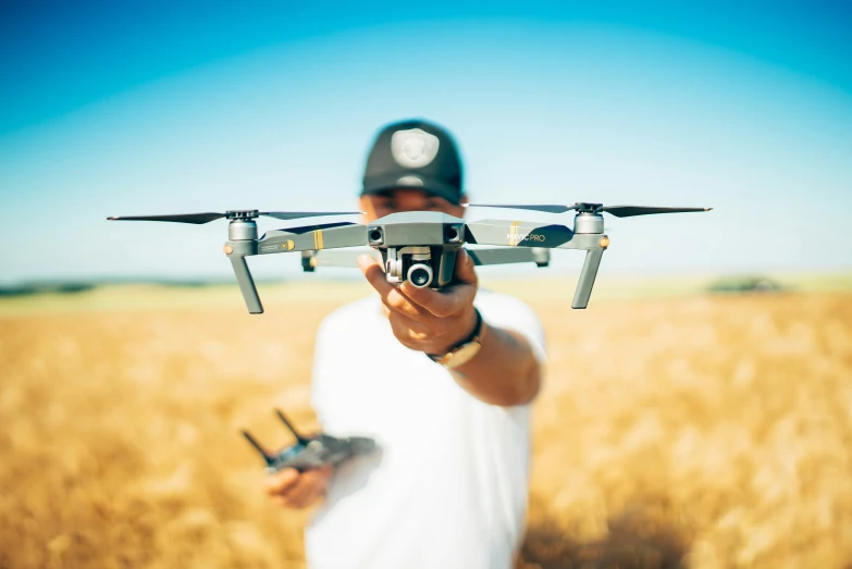 a man holding a black and white small camera flies a tiny white plane above a wheat field