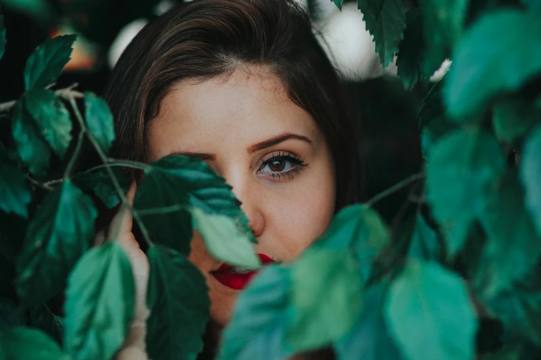 a close up of a woman peeking behind some green leaves