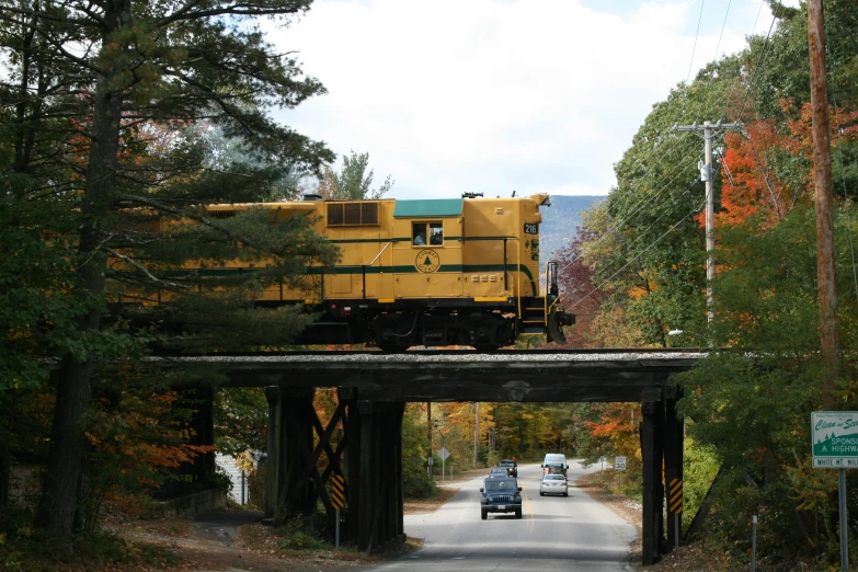 a yellow train engine is crossing an overpass near trees