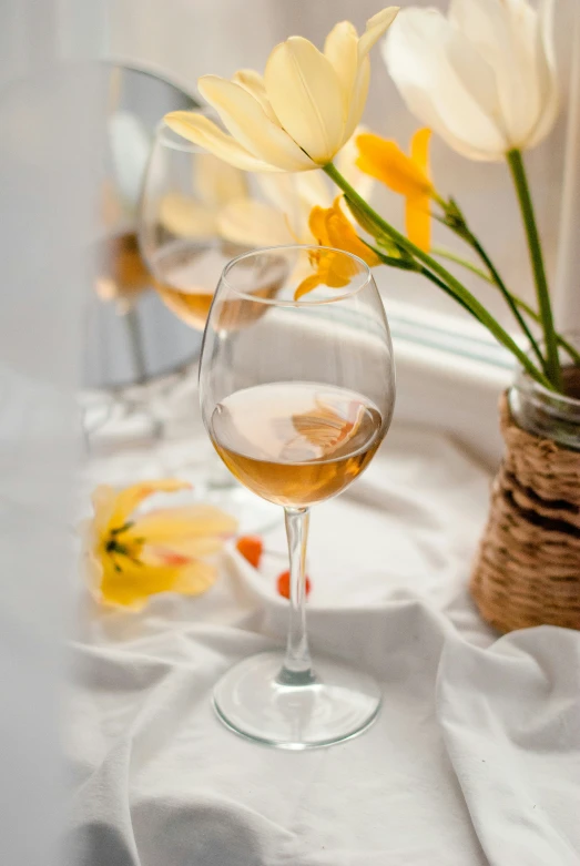 a wine glass with a small amount of white wine next to yellow flowers