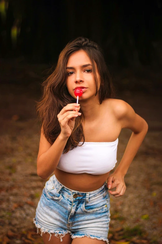 a beautiful woman in white shirt and jean shorts holding a lollipop
