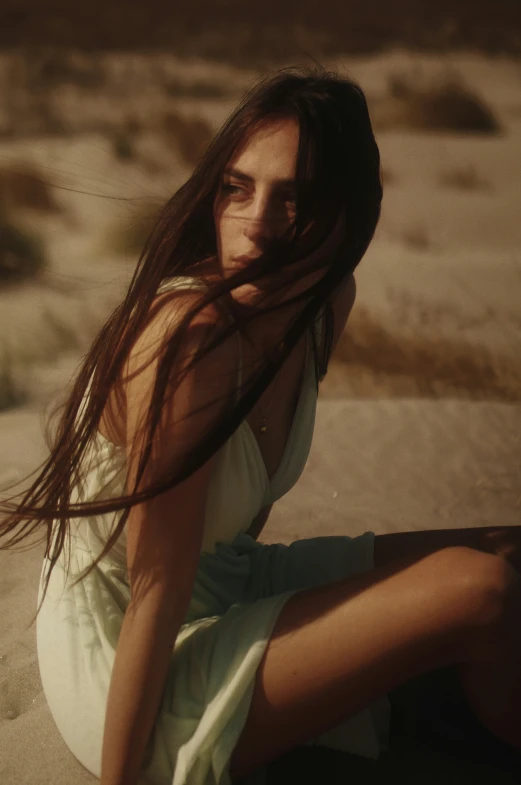 a woman sits in the sand wearing a white dress