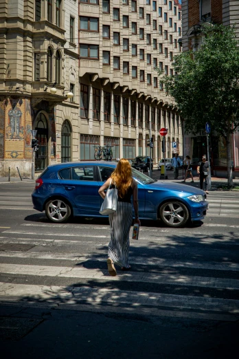 a woman walking across a street next to a blue car