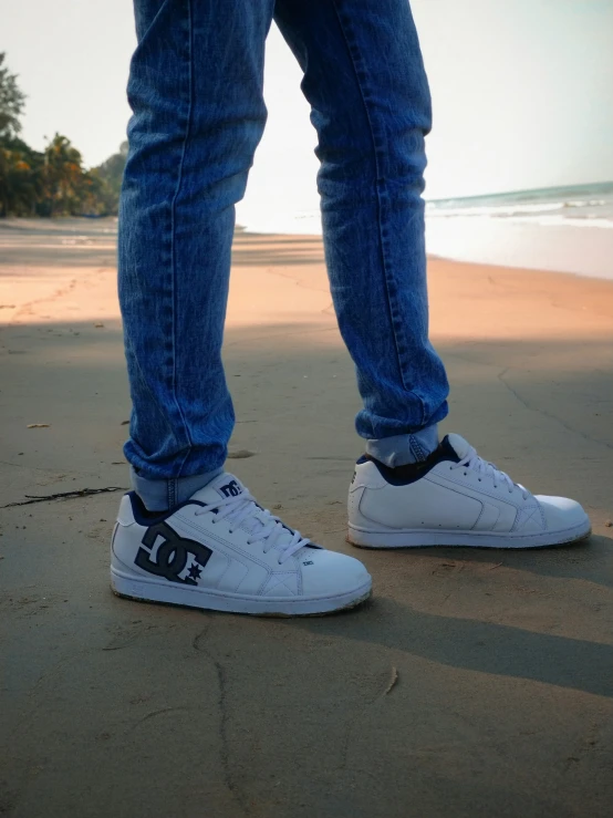 the legs of a person in white shoes standing near the ocean