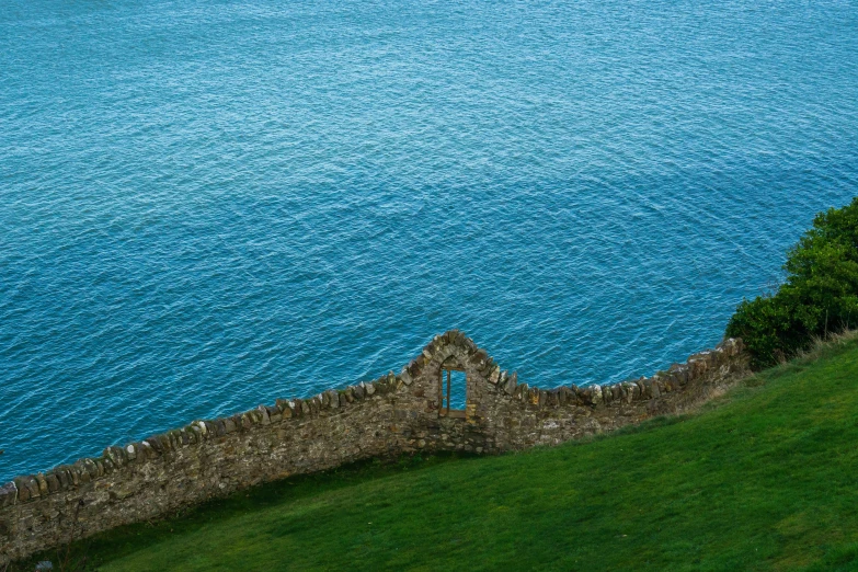 a stone structure sits on the edge of a grassy hill with blue water in the background
