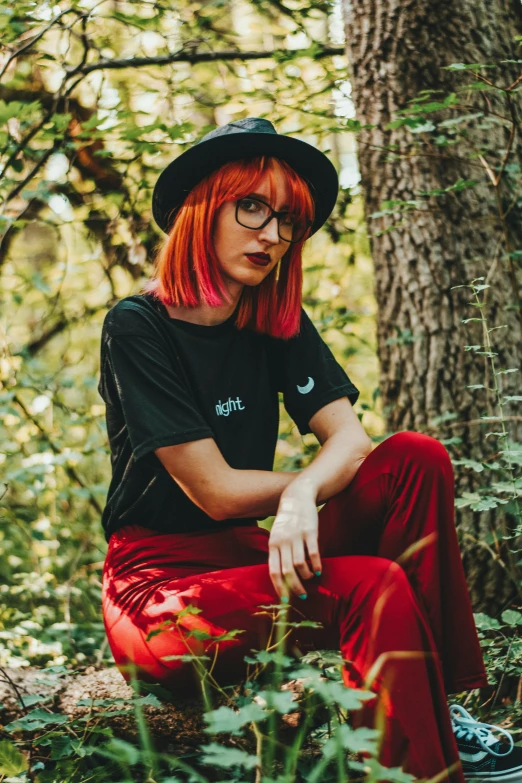 a woman with red hair wearing glasses, sitting on the ground in front of a tree