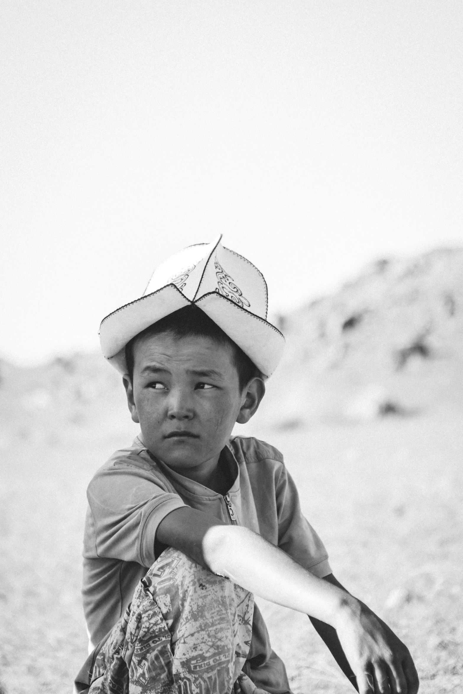 the boy is sitting on the dirt wearing a hat