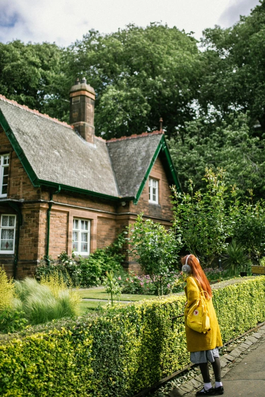 the woman is standing next to the hedge looking up at the house