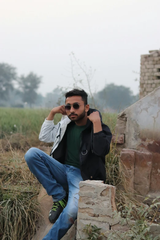 a man with glasses, jacket and sneakers sitting on a rock wall