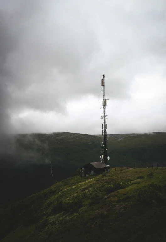 the cellular tower sits on a grassy hill under a cloudy sky