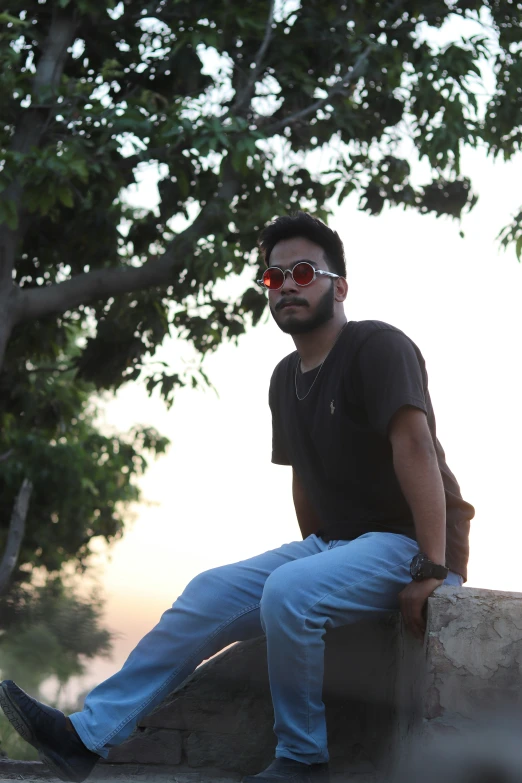 a man in black shirt and sunglasses sitting on ledge next to tree